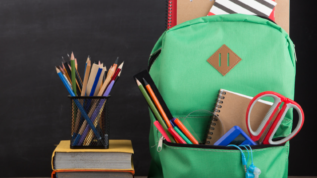 A green backpack filled with school supplies like notebooks, scissors and pencils sitting next to a stack of books with a pencil cup on top that is filled with colored pencils. A chalkboard is in the background. 