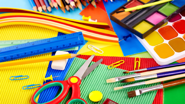 School supplies like scissors, a blue ruler, paintbrushes, watercolor paints, colored pencils, and paperclips lying on top of sheets of blue, green, red, and yellow construction paper. 