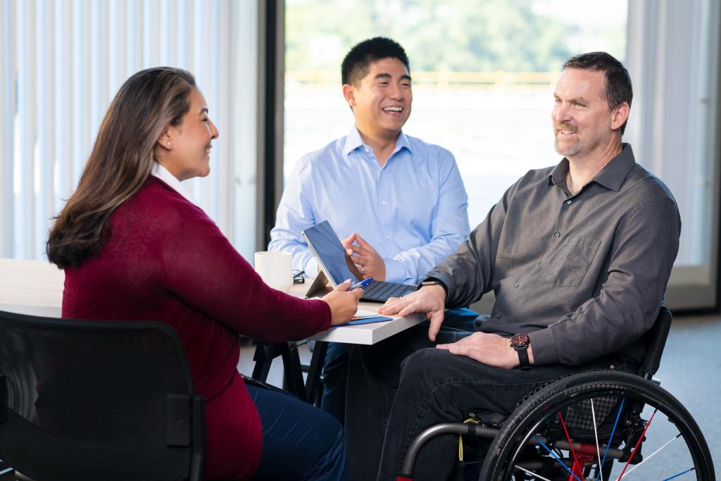 Three LADWP employees sitting and smiling around a table. The woman is wearing a red cardigan, white button-up shirt and blue pants and has medium length brown hair. One of the men is wearing a light blue button-up, navy pants, and has short black hair. The other man is wearing a grey button-up, a watch, black jeans, has short brown hair, and is in a wheelchair.