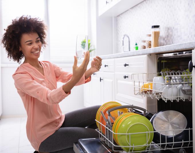 A woman with curly hair wearing a pink long-sleeve V-neck shirt is smiling at a clean glass that she is holding up. She is kneeling next to her dishwasher full of clean dishes.