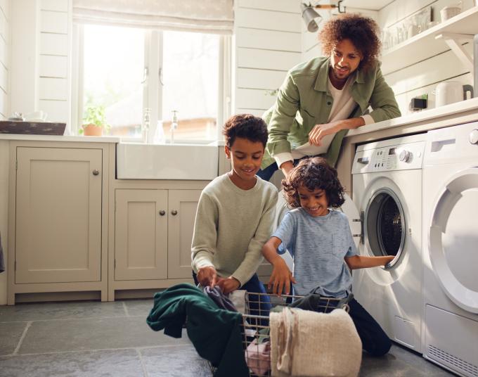 A man with curly hair is smiling and leaning against a white laundry machine, looking down at two kids. The two kids are smiling and taking things out of the laundry and putting it in a wire basket.