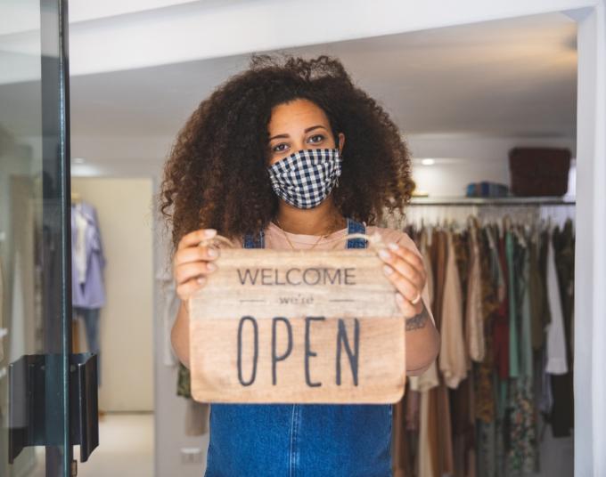 A shop owner wearing a face mask holds a sign in her business’s doorway that says: Welcome, we’re open.
