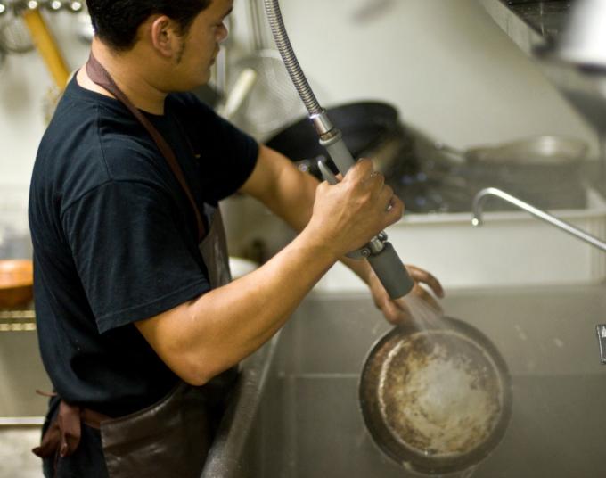A man wearing a black t-shirt and an apron uses a spray valve to rinse off a pan in a commercial kitchen’s sink.