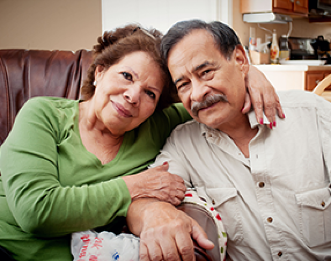 Two older adults sitting on a couch together. The woman has one arm around the man and the other arm holding his bicep. 