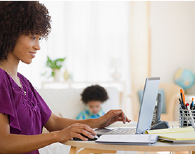 Smiling lady is sitting at a desk working on a laptop and a toddler is in the background.