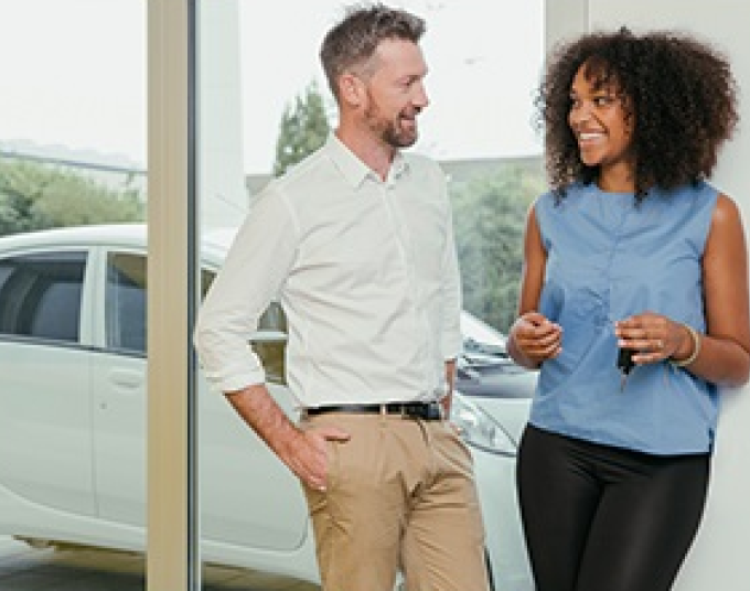 Man talking to a woman who is holding car keys and smiling. A car is parked behind them.