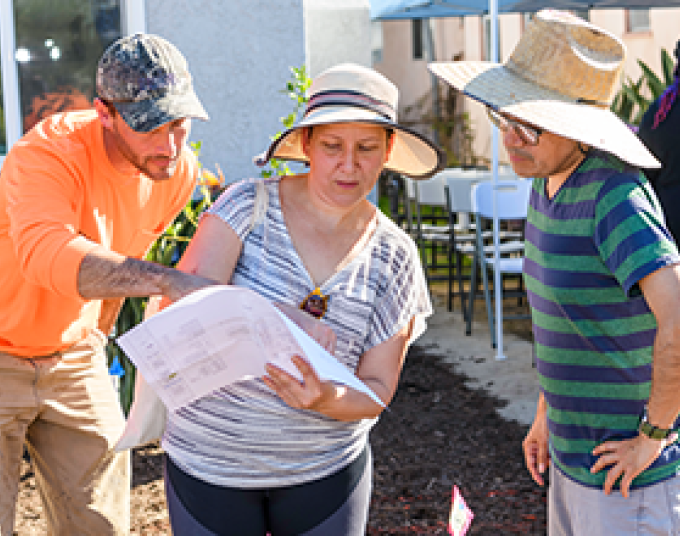 Three people wearing hats in the sun standing on dirt. The middle person is holding and looking at a large piece of paper with a design plan. The left person is pointing to something on the paper. The right person has a hand on their hip and is looking at the paper.