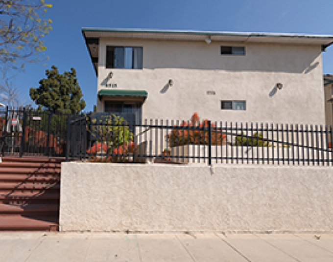 Beige apartment building on a sunny day in Los Angeles. In front of it is a black metal fence and to the left is an adobe colored staircase.