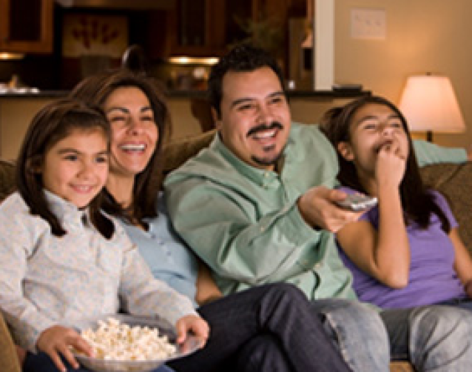 Family of four smiling and sitting on couch together while watching tv and eating popcorn. In the background there is a lamp on. 