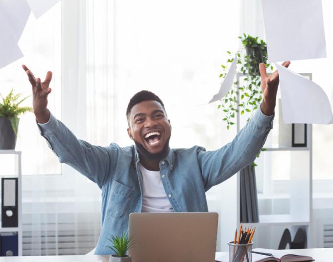 Smiling man throwing papers in the air with a laptop in front of him. 