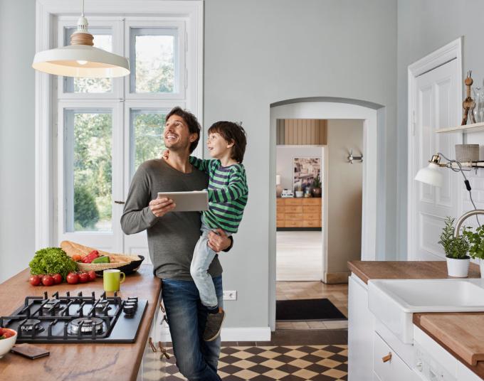 Smiling man carrying smiling kid while holding a tablet and looking up at the kitchen light. 