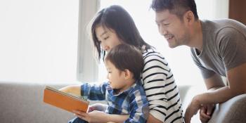 Man leaning over the shoulder of a woman seated with a child on her lap. She and the child are looking at a book.